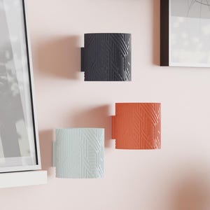 May include: Three wall-mounted, cylindrical sconces in a modern style. One is dark gray, one is light blue, and one is orange. Each has a textured, geometric pattern. The sconces are mounted on a pink wall.