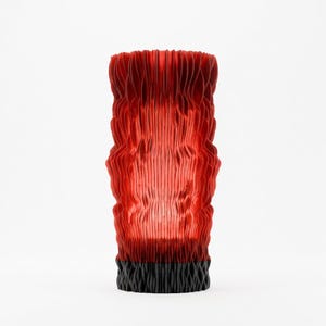 May include: A cylindrical vase featuring a vertical, pleated, and wavy texture. The upper portion is a vibrant, translucent red, while the base is a solid, opaque black. The structure consists of numerous thin, upright fins that create a rhythmic, rippled pattern throughout the entire height of the object.