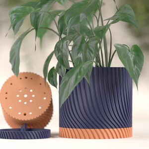 May include: A navy blue and orange plant pot with a matching saucer and a stackable lid. The pot has a ribbed design and contains a green leafy plant. The pot is approximately 10 inches tall.