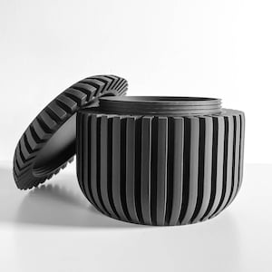 May include: A black, cylindrical container with a matching lid. The container has vertical grooves, and the lid has a ridged pattern. The lid is partially open, revealing the interior of the container. The design is modern and minimalist.