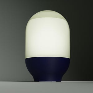 May include: A cylindrical table lamp with a rounded top and a smooth, matte finish. The upper portion is a translucent white shade that emits a soft glow, while the lower base is a solid, deep navy blue color with a curved, tapered shape.