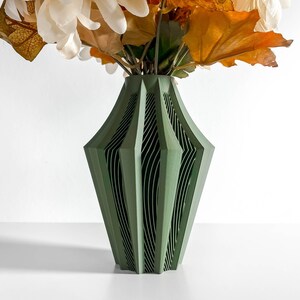 May include: A green, geometric vase with a unique, layered design. The vase holds a bouquet of white and orange artificial flowers and leaves. The vase is made of a solid material and has a modern aesthetic.