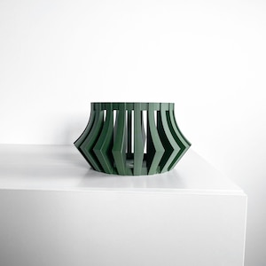 May include: A dark green decorative bowl with a geometric design. The bowl has a wide, open top and a base that tapers inward. The sides are made up of vertical and angled lines, creating a unique and modern look. The bowl is sitting on a white surface.