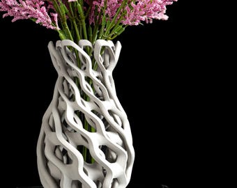 May include: A white 3D-printed vase with an intricate, open-lattice, wavy structure. The vase features a narrow neck and a wider, rounded body, containing a dense arrangement of bright pink astilbe flowers with green stems.