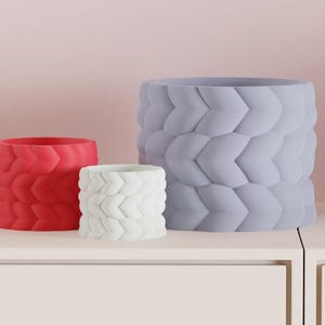 May include: Three decorative planters in a row, each with a textured, woven design. The planters are in three different colors: red, white, and light purple. The planters are cylindrical and sit on a white surface.
