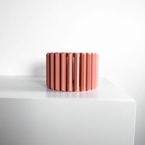 May include: A coral-colored, cylindrical decorative object with vertical, rectangular slats. The object is placed on a white surface against a white background. The design features a series of evenly spaced, vertical elements, creating a textured appearance.