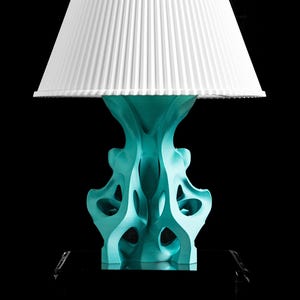 May include: A table lamp featuring a white, pleated, conical lampshade atop a turquoise, organic, lattice-structured base. The base has a complex, hollow, branching design with multiple openings, resting on a dark, reflective square platform.