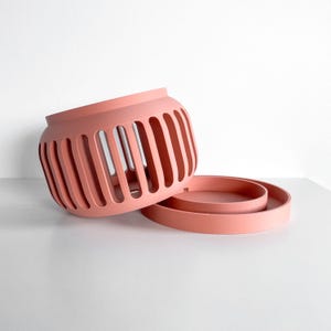 May include: A coral-colored, slotted container with a matching circular base. The container has a cylindrical shape with vertical slots. The base is a flat, round disc. The objects are on a white surface.