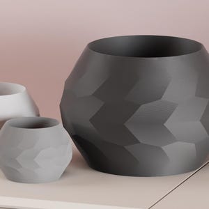 May include: Three geometric planters in shades of gray and white. The largest planter is dark gray, the medium one is light gray, and the smallest is white. The planters have a faceted design and are arranged on a light-colored surface.