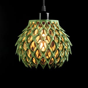 May include: A sage green pendant light fixture featuring a complex, lattice-like geometric structure with pointed, leaf-like protrusions. The shade surrounds a central light bulb that emits a warm, golden glow, suspended by a black cord.