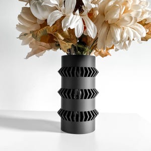May include: A black cylindrical vase with three decorative bands of arrow-shaped cutouts. The vase holds a bouquet of cream and tan-colored artificial flowers. The vase is made of a matte material and sits on a white surface.