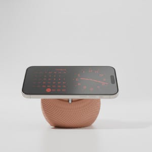 May include: A smartphone resting horizontally on a peach-colored, spherical stand with a textured, dimpled surface. The phone screen displays a digital calendar for August and an analog clock face with red numerals and hands against a dark background.