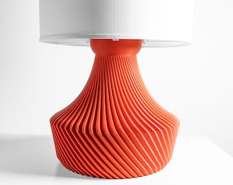 May include: A table lamp featuring a vibrant orange base with a distinct spiraling, ribbed texture that tapers toward the top. The lamp is topped with a plain, cylindrical white lampshade that provides a neutral contrast to the textured base.