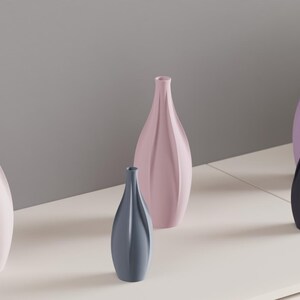 May include: A collection of decorative vases in various pastel colors, including light pink, lavender, and gray-blue. The vases have a slender, elongated shape with a ribbed texture, arranged on a white surface against a gray background.