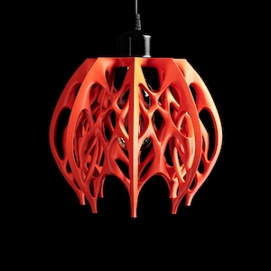 May include: A modern, orange pendant light fixture with a unique, openwork design. The light has a spherical shape with a complex, web-like pattern, suspended from a black cord against a black background. The fixture's intricate design allows light to filter through.