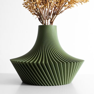 May include: A green decorative vase with a unique, geometric design. The vase has a wide base that tapers to a narrower neck. The vase is filled with dried, tan-colored flowers. The vase is made of a ridged material.