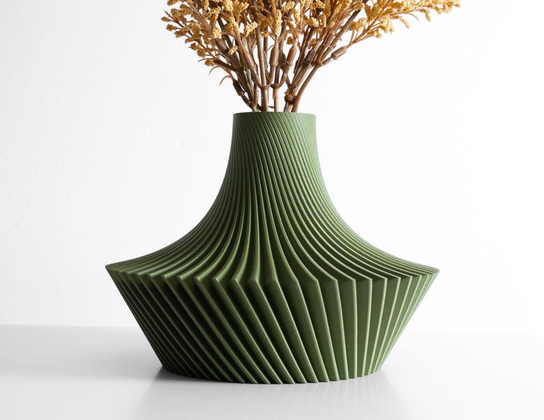 May include: A green decorative vase with a unique, geometric design. The vase has a wide base that tapers to a narrower neck. The vase is filled with dried, tan-colored flowers. The vase is made of a ridged material.
