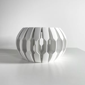 May include: A white, geometric decorative bowl with a rounded shape. The bowl features a series of vertical, angular cutouts that create a pattern of light and shadow. The design is modern and minimalist, suitable for home decor.