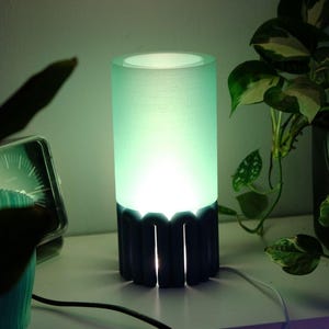 May include: A cylindrical table lamp with a green glow. The lamp has a dark base with vertical cutouts and a light green shade. A white cord extends from the base. Potted plants are in the background.