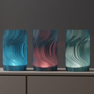 May include: Three cylindrical decorative lamps with a wavy, textured design. The lamps are lit from within, showcasing blue, red, and green hues. They sit on a neutral-toned surface, creating a modern and artistic display.