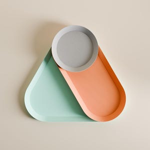 May include: A set of three nested trays featuring a triangular mint green tray, an elongated oval orange tray, and a circular light gray tray. The trays are arranged in an overlapping formation, creating a geometric composition with distinct colors.