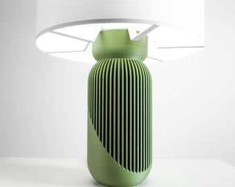 May include: A table lamp featuring a cylindrical sage green base with vertical ribbed detailing and a diagonal cut-out design. The lamp is topped with a wide, white drum-shaped lampshade that emits a bright, diffused light from within.