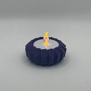 May include: A purple, geometric-patterned candle holder with a white interior holds a small, battery-operated LED candle. The LED candle emits a warm, flickering light, resembling a real flame. The candle holder is a decorative item.