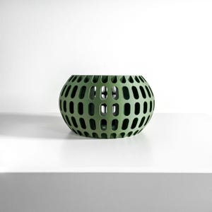 May include: A spherical olive green bowl featuring a repeating pattern of vertical oval cutouts across its entire surface. The object has a wide, open top and a rounded base, creating a hollow, cage-like structure with a matte finish throughout.