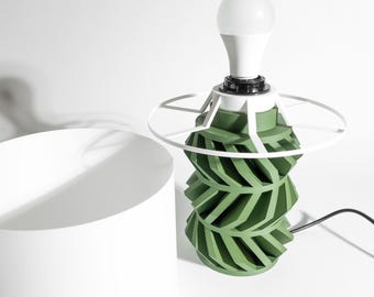 May include: A table lamp featuring a cylindrical base with a repeating green chevron pattern, topped with a white circular frame and a white light bulb. A separate, plain white cylindrical lampshade sits to the left of the lamp base.