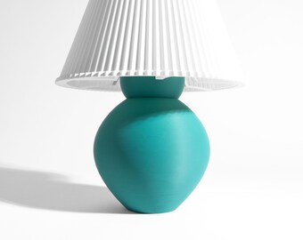 May include: A table lamp featuring a rounded, teal-colored base with a smooth, matte finish. The lamp is topped with a white, conical lampshade that has a distinct, vertical pleated texture throughout its entire surface area.