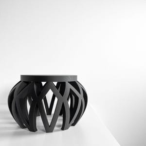 May include: A black, modern side table with a geometric design. The table has a round top and a base with a series of intersecting, curved lines. The table is made of a solid, dark material and sits on a white surface.