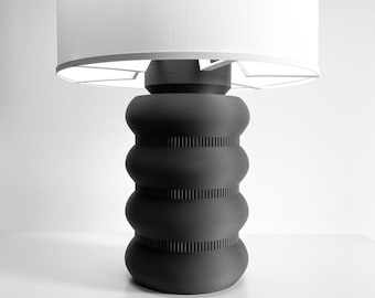 May include: A table lamp featuring a matte black base with a stacked, rounded, ribbed design and a wide, cylindrical white lampshade. The base consists of four distinct bulbous sections separated by narrow, textured bands that provide visual contrast.