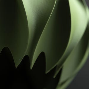 May include: A matte olive green object with a curved, spiraling, and layered structure. The surface features smooth, sweeping ridges that create deep shadows and sharp, pointed edges along the bottom, forming a complex, three-dimensional shape.