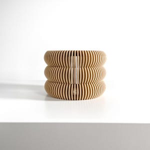 May include: A light brown, cylindrical wooden planter with a layered, ribbed design. The planter has three stacked sections, each with horizontal grooves, creating a textured surface. A vertical support runs through the center.