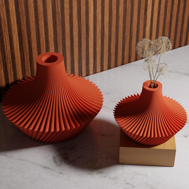 May include: Two orange, spiral-shaped vases. One vase is larger and sits on a marble surface, while the other, smaller vase rests on a wooden block. The smaller vase holds dried flowers. The background features a wooden panel.