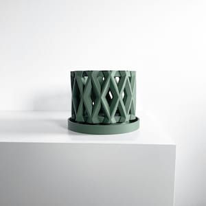 May include: A sage green cylindrical planter featuring a geometric lattice pattern with diamond-shaped cutouts. The planter sits on a matching circular base tray, creating a cohesive unit with a matte finish and a structured, open-work design.