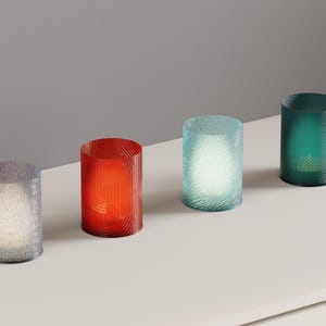 May include: Four cylindrical glass candle holders arranged in a row. The holders are translucent in shades of grey, red, light blue, and dark green, each featuring a textured, rippled surface pattern that catches the light from within.