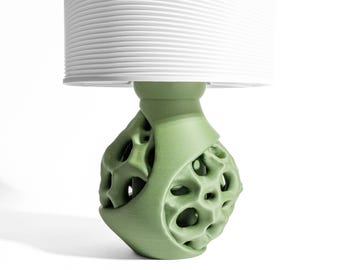 May include: A table lamp featuring a sage green base with an organic, porous, and textured surface design. The lamp is topped with a cylindrical white lampshade that has fine horizontal ribbed detailing around its entire exterior circumference.