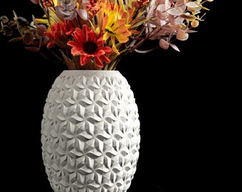 May include: A white cylindrical vase with a repeating geometric star pattern on the exterior, filled with an arrangement of artificial flowers in shades of red, orange, and yellow, including berries and foliage against a black background.