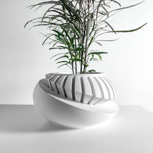 May include: A modern white planter with a unique, layered design. The top section features vertical, angled cutouts, while the base has a smooth, curved shape. The planter holds a green leafy plant, creating a striking contrast.