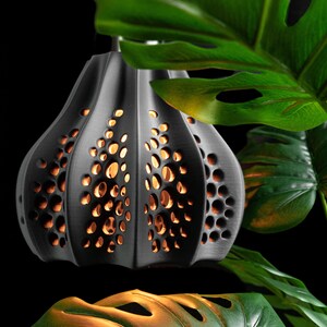 May include: A matte black pendant light fixture with a bulbous, ribbed shape and a series of irregular, organic-shaped cutouts. Warm, golden light glows from within the dark structure, illuminating the interior through the perforated pattern.