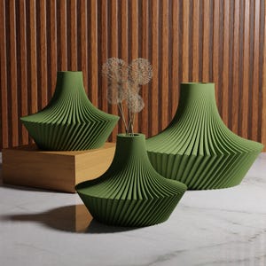 May include: Three green decorative vases with a unique, ribbed design. The vases vary in size and shape, with a wide base and a narrower neck. One vase holds dried flowers. The background features a wooden wall and a marble surface.