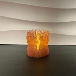May include: A cylindrical candle holder with a translucent orange, petal-like geometric pattern. The structure features a repeating vertical design with pointed edges at the top rim, emitting a warm, glowing light from within the center.