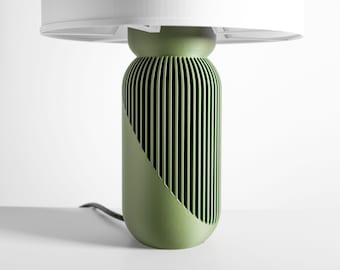 May include: A table lamp with a cylindrical olive green base featuring vertical slats and a diagonal cutaway design. The lamp is topped with a wide, white circular lampshade. A black power cord extends from the base of the lamp.