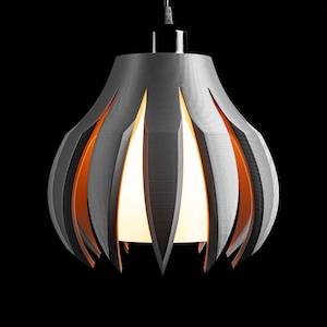 May include: A pendant light fixture featuring a dark gray, petal-shaped outer shade with pointed edges. The shade surrounds a central, glowing white inner light source that emits a warm orange light from the gaps between the gray petals.
