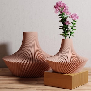May include: Two decorative, light-pink vases with a unique, ridged design. One vase holds pink flowers with green stems. The vases are displayed on a wooden surface and a wooden block. The vases have a wide base and a narrow neck.