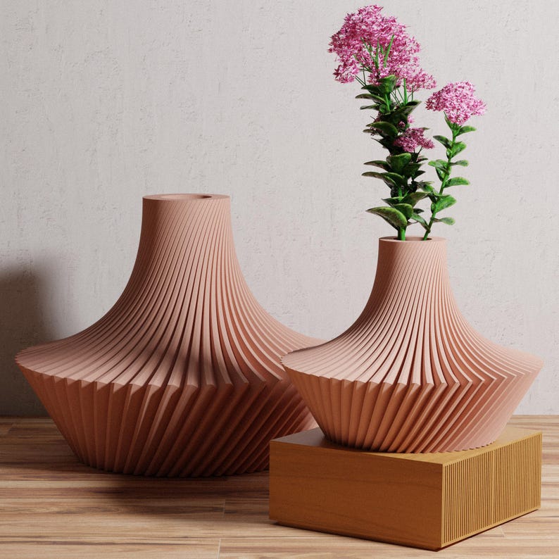 May include: Two decorative, light-pink vases with a unique, ridged design. One vase holds pink flowers with green stems. The vases are displayed on a wooden surface and a wooden block. The vases have a wide base and a narrow neck.