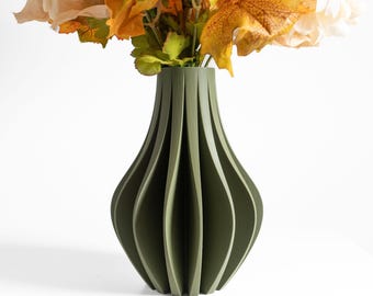 May include: An olive green vase with a vertical ribbed, fluted design and a narrow neck. The vase is filled with a bouquet of artificial flowers featuring cream-colored petals and orange, yellow, and green autumn-toned maple leaves.