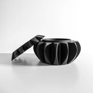 May include: A matte black circular container with a removable lid. The exterior features a series of vertical, curved fins that create a repeating pattern around the body. The lid has a matching geometric, angular edge design.