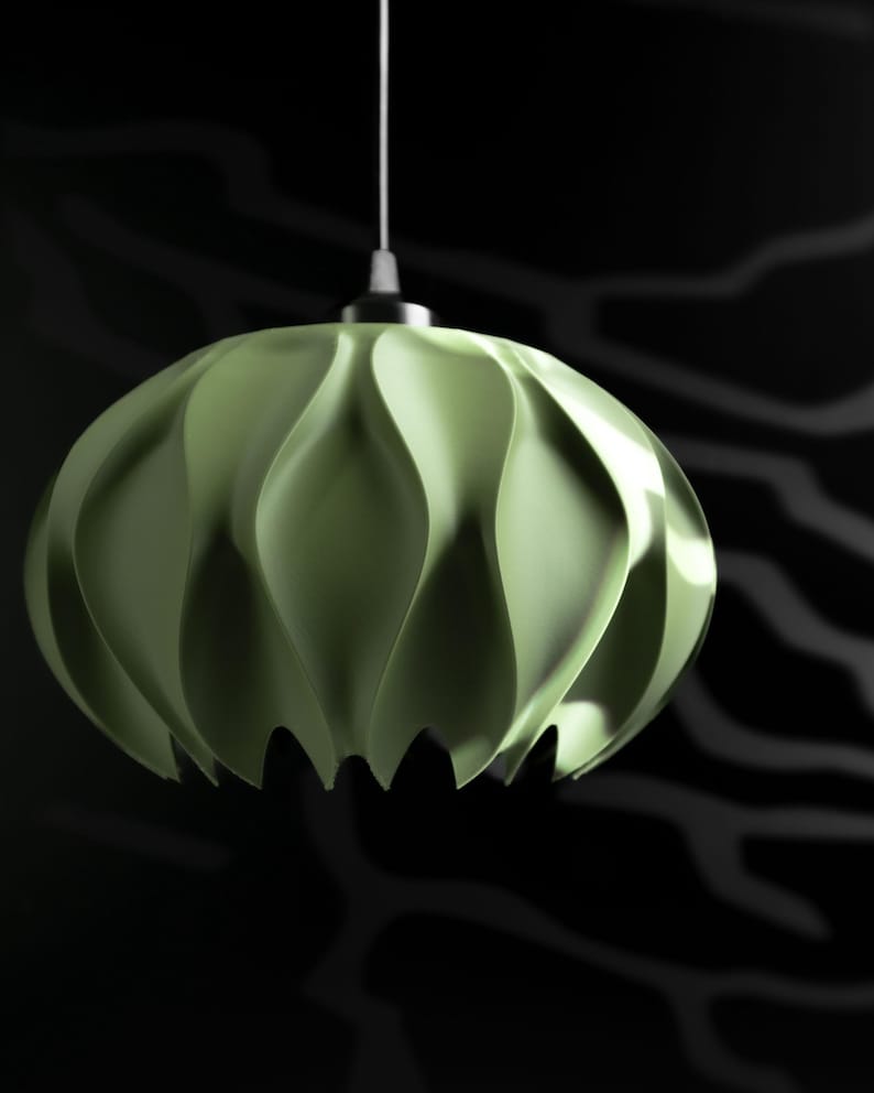 May include: A pale green pendant light fixture with a rounded, multi-faceted shape featuring deep, vertical, wave-like folds. The lamp is suspended by a thin white cord attached to a metallic silver cap at the top center of the structure.